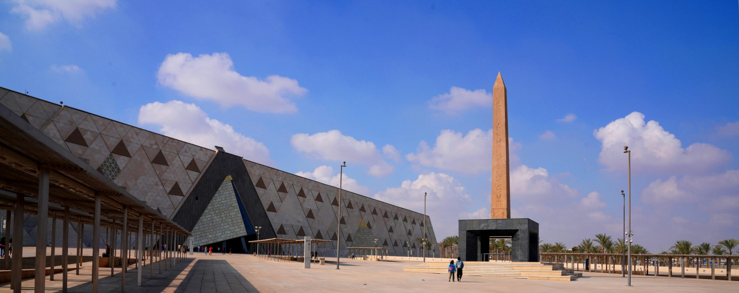 Exterior View of the Grand Egyptian Museum with its modern architecture , wide plaza and the tall Ramses II obelisk.