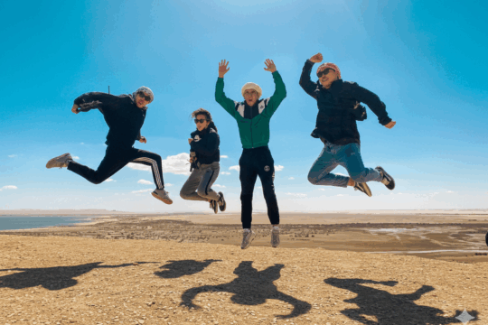 Four friends joyfully jumping against a bright blue sky on a desert hilltop, overlooking a vast landscape. Keywords: adventure travel, group tours, holiday vacation, desert landscape, joyful moment, travel photography.
