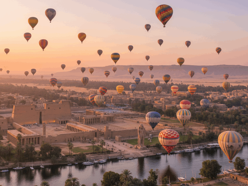 https://holidaytours.one/14-days-egypt-tour-package/ Hot air balloons flying over Luxor at sunrise, overlooking ancient temples along the Nile River and the West Bank of Egypt